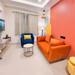 Modern living room with orange sofa, blue armchair, wall-mounted TV, and stylish decor in a fully furnished service apartment for corporates