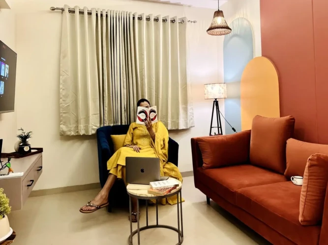 Woman reading book while sitting on a modern blue armchair in a fully furnished living room of service apartment for coporates